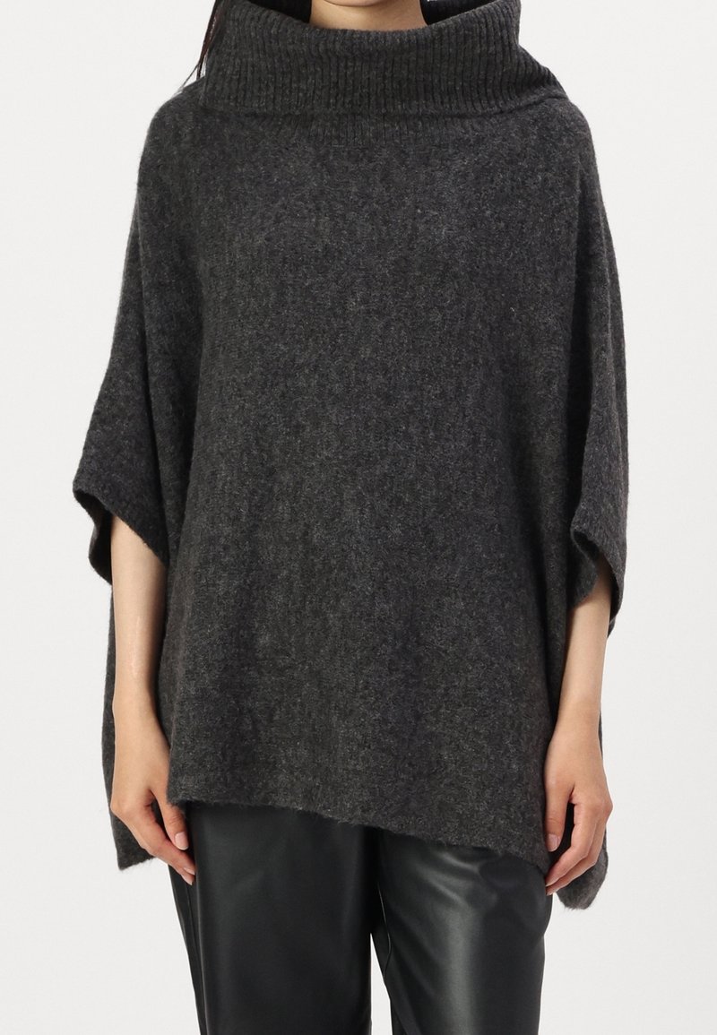 Personne portant un poncho tricoté ample gris foncé avec un col côtelé haut et des manches courtes sur un pantalon noir.