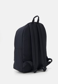Mochila de tela negra con respaldo acolchado, dos correas ajustables y un asa en la parte superior. Textura suave, forma redondeada y diseño minimalista.