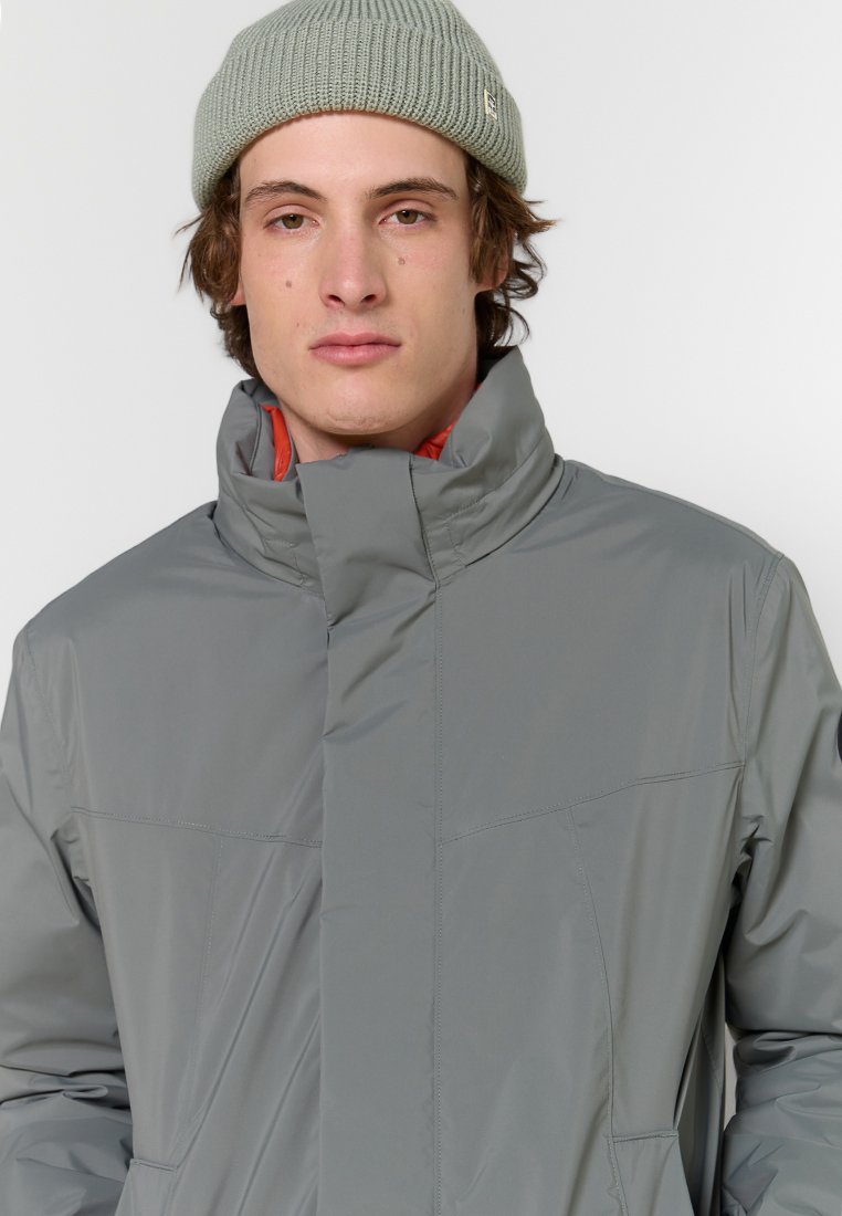 Giovane uomo che indossa un giubbotto invernale grigio chiaro con colletto alto e un berretto di maglia verde chiaro, che guarda dritto davanti a sé.