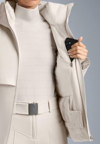 Bež jakna otporna na vodu s visokim ovratnikom, zatvorenim džepom i rebrastim bijelim gornjim dijelom. Dovršena teksturiranim remenom i elegantnim šavovima.