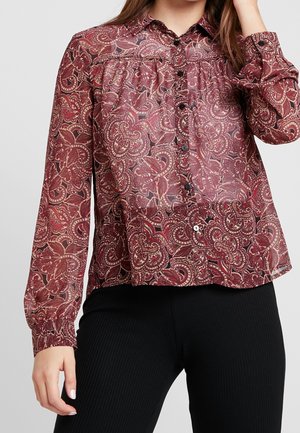 Kvinde iført langærmet knapbluse med burgundy paisleymønster og sorte ribbukser, hånd hævet til hovedet.