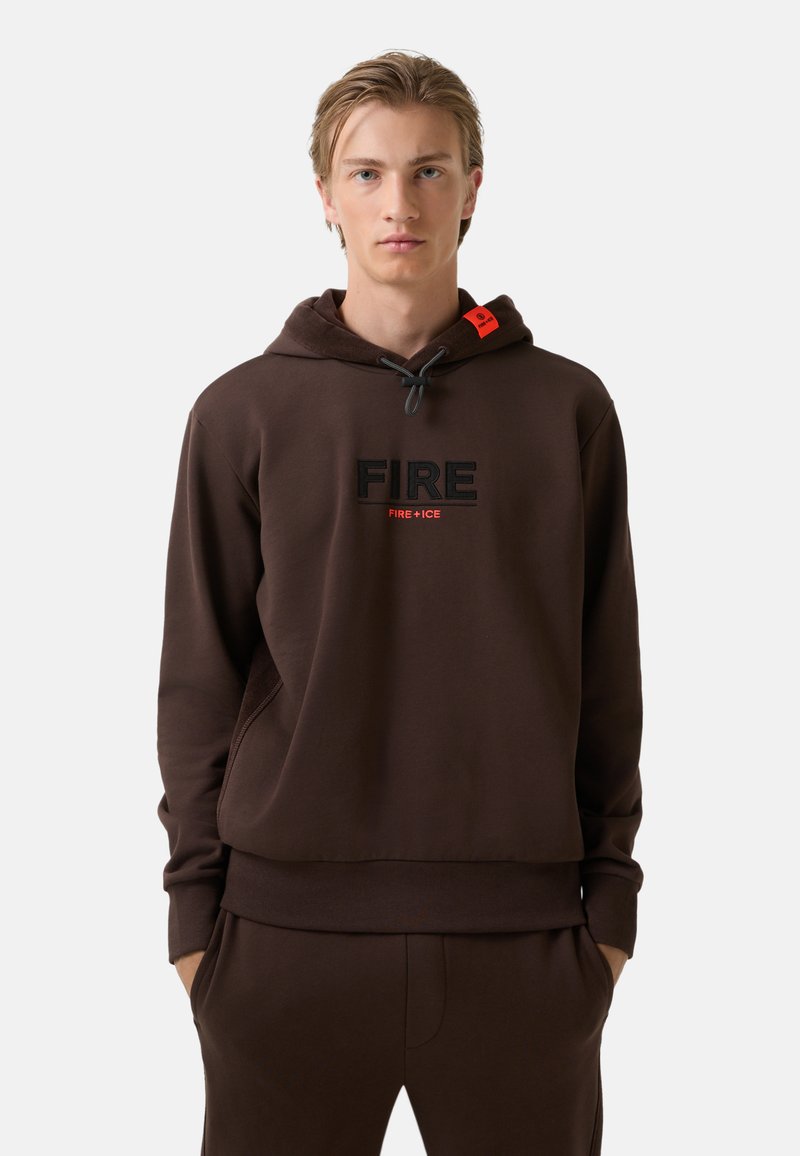 Młody mężczyzna w ciemnobrązowym hoodie z napisem "FIRE" oraz "FIRE+ICE" na piersi, z rękami w kieszeniach, na neutralnym tle.