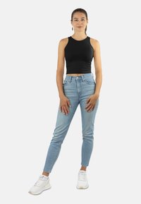 Frau steht mit einem schwarzen ärmellosen Crop-Top, hellblauen hochgeschnittenen Jeans, weißen Sneakern und hängenden Ohrringen vor einem weißen Hintergrund.