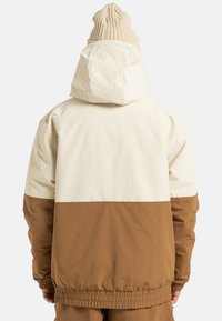 Kurtka z kapturem o dwutonowym wzorze; górna część w kolorze kremowym, a dolna w brązowym, wykonana z gładkiego, lekkiego materiału. Elastyczne mankiety i dół.