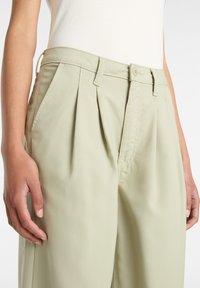 Pantaloni plissettati verde chiaro con vita aderente, chiusura con bottone e tasche laterali. Il materiale appare liscio con una texture minima.