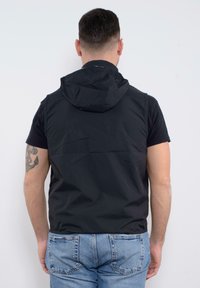 Gilet senza maniche nero con cappuccio, realizzato in un tessuto leggero e liscio, con cuciture in vista, indossato sopra una maglietta nera e jeans blu.