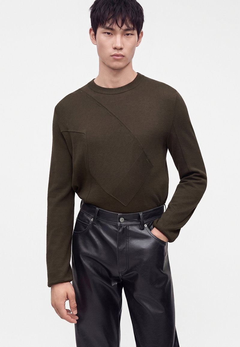 Young man wearing a dark olive green sweater with textured patterns and black leather pants, standing with one hand in pocket.