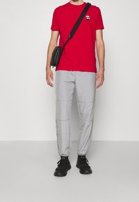 Červené bavlněné tričko s malou černou grafikou lebky, spárované se světle šedými, lehkými, zipovými kalhotami a černými teniskami.