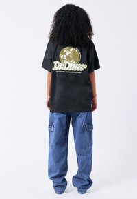 Černé oversize tričko s kulatým grafickým logem v olivové barvě na zadní straně, v kombinaci s volnými modrými džínovými kalhotami cargo s bočními kapsami.