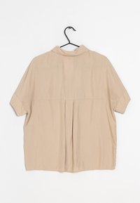 Beige short-sleeve shirt with a collar, featuring a back pleat and smooth texture, hanging on a black plastic hook against a white backdrop.