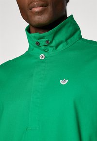 Moški oblečen v zeleno majico s kragnico, ki ima tri gumbe in majhen bel in moder logotip na prsih.