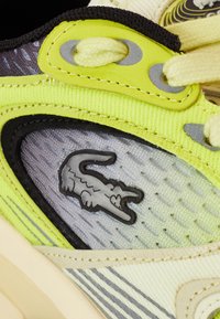 Tenisky Lacoste kombinující žluté, šedé a černé materiály. Mají texturovaný design a výrazné logo Lacoste v šedé barvě.