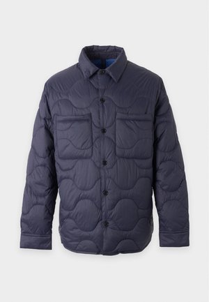 Mann trägt eine marineblaue gesteppte Jacke über einem grün gestreiften Hemd, hellblaue Jeans und weiße Turnschuhe und steht vor einem schlichten weißen Hintergrund.