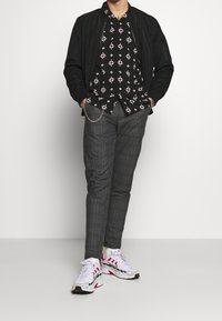 Černá bombera přes černé tričko s bílým květinovým vzorem, šedé kárované kalhoty a bílé tenisky s červenými akcenty a stříbrnými detaily.