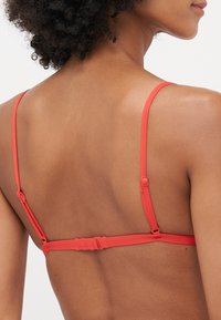 Rød bralette med tynne justerbare stropper, en krok-lukking bak og en glatt tekstur. Minimalistisk design med en enkel form.