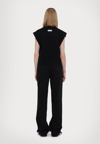 Czarny bezrękawnik z wysokim kołnierzem, z etykietą z tyłu. Nosi się go z czarnymi spodniami o szerokich nogawkach i teksturowanym obuwiem.