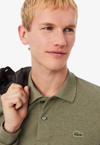 Camisa polo verde con un acabado texturizado, dos botones y un pequeño logo de cocodrilo bordado en el pecho; modelo sosteniendo una chaqueta negra.