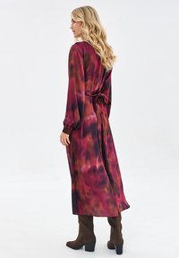 Długa, zwiewna sukienka z długim rękawem w czerwono-brązowy wzór tie-dye z paskiem w talii, wykonana z gładkiego materiału i z lekko bufkowymi rękawami.