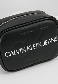 Czarna skórzana torba z wytłoczonym logo "CK" oraz "CALVIN KLEIN JEANS" w kolorze białym. Posiada zamek błyskawiczny i prostokątny kształt. Gładka faktura.