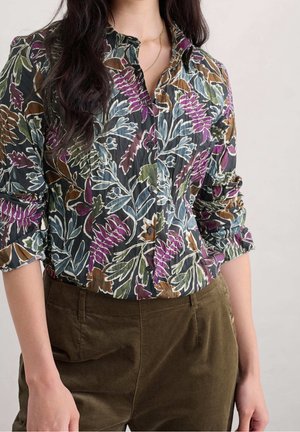 Femme portant une chemise fleurie boutonnée avec des feuilles violettes, vertes et brunes, associée à un pantalon marron, sur un fond uni clair.