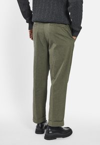 Pantalon en laine vert olive à finition texturée, doté de poches latérales, d'une coupe décontractée et de revers retroussés, associé à des chaussures noires.