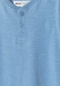 Šviesiai mėlynas, tekstūruotas trumpas rankovėmis marškinėliai su trimis mygtukais prie apykaklės ir išsiuvinėjimu kairėje pusėje.