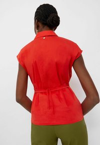 Vrouw met gevlochten haar in een rode mouwloze top met tailleband, met haar rug naar ons gekeerd, in olijfgroene broek tegenover een effen achtergrond.