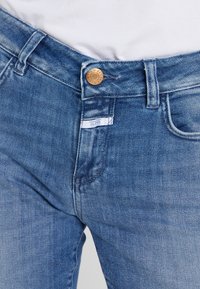 Jeans de denim azul con un corte clásico, botón prominente en la cintura y una pequeña etiqueta de la marca cerca del bolsillo. Textura suave con ligero desvanecimiento.