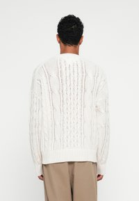 Fehér kötött pulóver, amely kábelmintával, bordás szegéllyel és laza fazonnal rendelkezik. A hátsó részén bonyolult textúra és nyitott kötött minta látható.