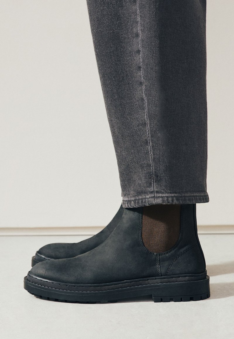 Schwarze Chelsea-Stiefel aus Veloursleder, getragen mit dunkelgrauen Jeans mit geradem Bein auf hellem Boden und neutralem Wandhintergrund.