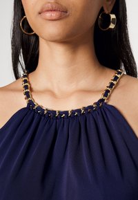 Kvinna som bär en marinblå halterneck-top med guldkedjespetsar och stora guldörhängen, där endast halslinjen och läpparna syns.