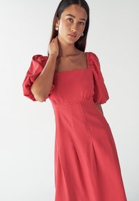 Rdeča obleka z napihnjenimi rokavi, kvadratnim izrezom, oprijetim korpusom in padajočo krilo, izdelana iz mehkega materiala z diskretnim teksturo.