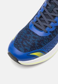 Zila sporta kurpe ar tekstūru auduma virsmu, sietiņš paneļiem, kontrastējošiem dzelteniem akcentiem un mīkstu baltu zoli.