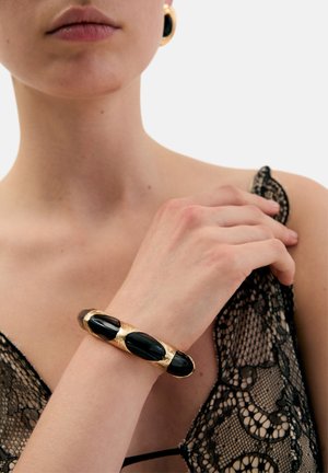 Kultainen ja musta rannerengas, jossa on sileitä mustia korostuksia ja teksturoituja kultaisia yksityiskohtia, kädessä, jossa on musta pitsinen vaatekappale.