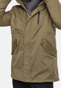 Olivgrüne Jacke mit Kapuze, ausgestattet mit zwei Fronttaschen, einem Reißverschluss und einem strukturierten grauen Schal unter dem Kragen.