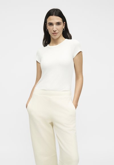 Γυναίκα με σκούρα μαλλιά φοράει εφαρμοστή λευκή μπλούζα με κοντά μανίκια και εκρού πλατιά παντελόνια με ψηλή μέση, με τα χέρια στις τσέπες, σε απλό φόντο.