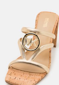 Sandale beige à talon bloc en liège avec des sangles en cuir doré présentant un logo circulaire doré MK sur la sangle supérieure.