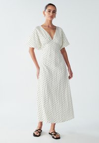 White maxi dress with black polka dots, deep V-neck, flared sleeves, textured fabric, featuring a flowing silhouette and minimalistic design.