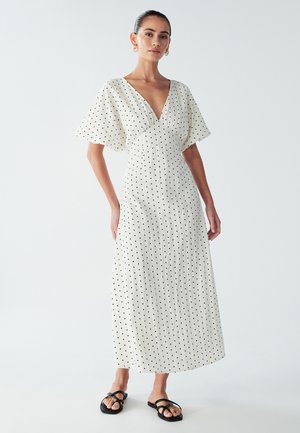 White maxi dress with black polka dots, deep V-neck, flared sleeves, textured fabric, featuring a flowing silhouette and minimalistic design.