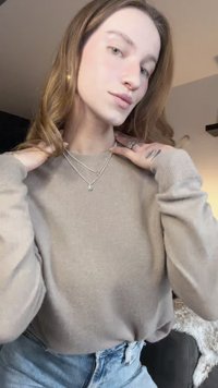 Béžový žebrovaný sveter s dlhými rukávmi, vrstvené strieborné náhrdelníky, modré džínsy s vysokým pásom a vlnité rozpustené vlasy.