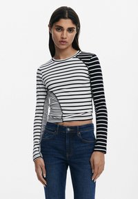 Schwarz-weiß gestreiftes langärmliges Crop-Top mit kontrastierenden Ärmeldesigns. Rippenstruktur, runder Ausschnitt, taillierte Passform. Kombiniert mit dunkelblauen Jeans.