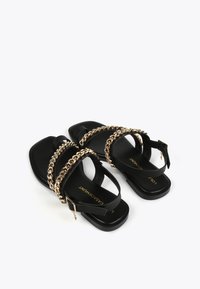 Mustat litteät sandaalit kultaketjuakselilla, joissa on sileä nahkainen yläosa, avoin varvasmuotoilu ja soljella varustettu hihna turvallista käyttöä varten.