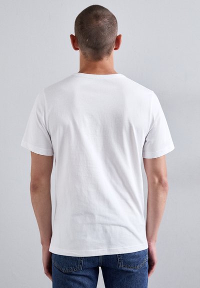Mann med kort hår iført en enkel hvit t-skjorte med korte ermer og blå jeans, stående mot en lys grå bakgrunn, med ryggen til betrakteren.