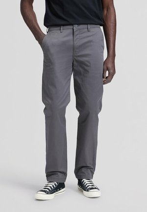Hombre con pantalones grises de pierna recta, camiseta negra y zapatillas deportivas blancas y negras, de pie con las manos en los bolsillos frente a un fondo liso.