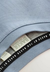 Lichtblauwe trui met een gestructureerd oppervlak, voorzien van een zwarte accentenband aan de binnenkant met witte tekst en twee merklables in contrasterende kleuren.