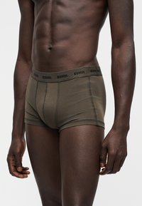 Buste masculin portant un boxer Diesel vert olive, montrant des abdominaux et des bras définis, sur un fond gris clair uni.