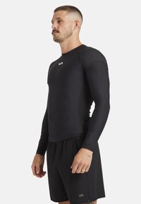 Zwart rash guard met lange mouwen, gemaakt van rekbaar materiaal. Strak model, gladde textuur, met een klein logo op de borst. Gecombineerd met zwarte shorts.