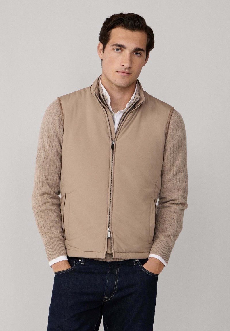 Jonge man in een beige mouwloze zip-up vest over een beige kabelgebreide trui en donkerblauwe spijkerbroek, staande met zijn handen in de zakken.