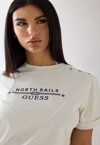 Femme aux cheveux lisses et foncés portant une chemise blanche North Sails Guess avec des détails de boutons sur l'épaule, regardant légèrement vers la gauche.