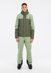 Grüne Skijacke mit einem dunkelgrünen Brustpanel, Kapuze und verstellbaren Bündchen, kombiniert mit hellgrünen Skihosen und schwarzen Skistiefeln.